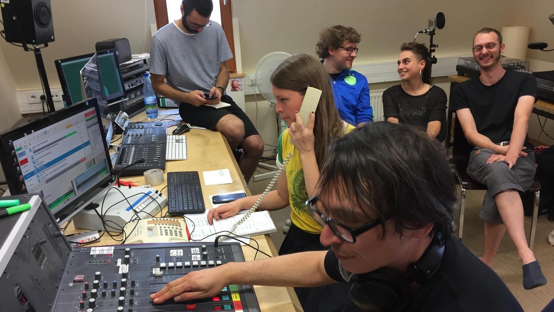Sechs junge Menschen sitzen in einem Radiostudio, ein Student bedient das Mischpult während eine Studentin telefoniert Sechs junge Menschen sitzen in einem Radiostudio, ein Student bedient das Mischpult während eine Studentin telefoniert