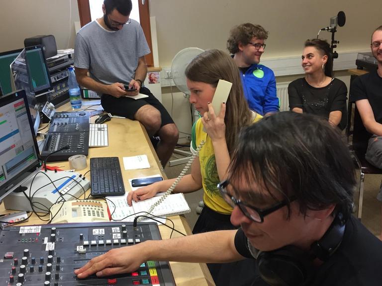 Sechs junge Menschen sitzen in einem Radiostudio, ein Student bedient das Mischpult während eine Studentin telefoniert