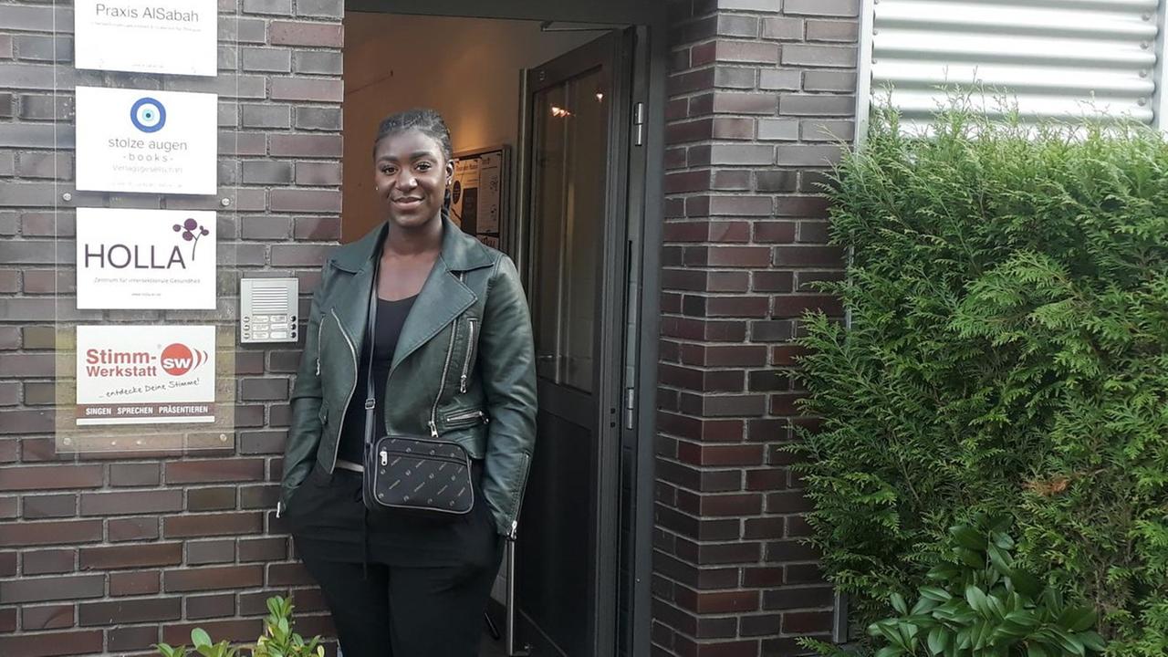 Eine junge schwarze Frau mit Lederjacke und schwarzer Hose steht vor einer Hauswand mit Briefkästen und Firmenschildern. Eine junge schwarze Frau mit Lederjacke und schwarzer Hose steht vor einer Hauswand mit Briefkästen und Firmenschildern.