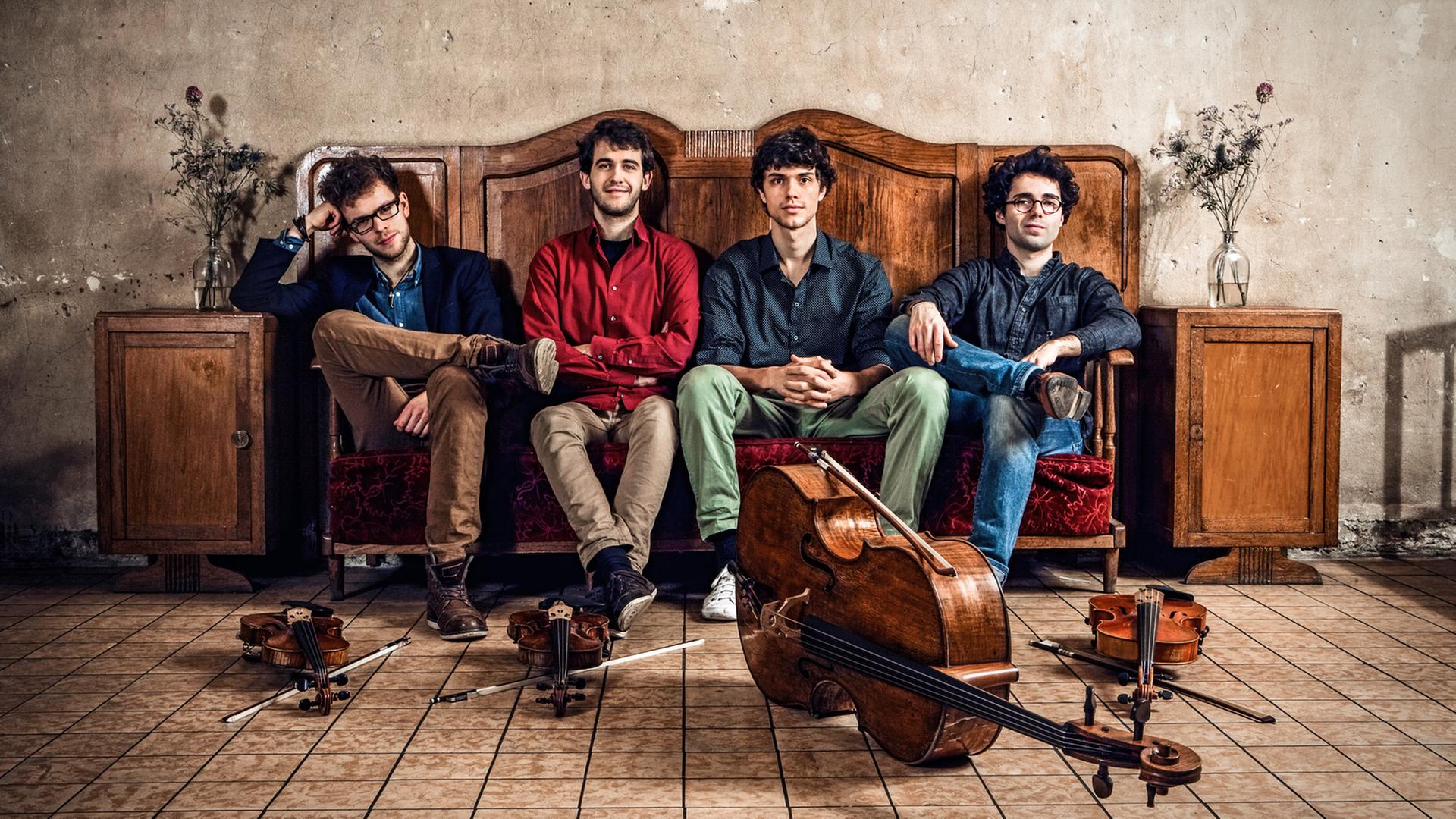 Vier junge Männer sitzen auf einem Vintagesofa aus dunkelbraunem Holz, vor ihnen liegen ihre Instrumente: zwei Violinen, ein Cello und eine Bratsche. Vier junge Männer sitzen auf einem Vintagesofa aus dunkelbraunem Holz, vor ihnen liegen ihre Instrumente: zwei Violinen, ein Cello und eine Bratsche.