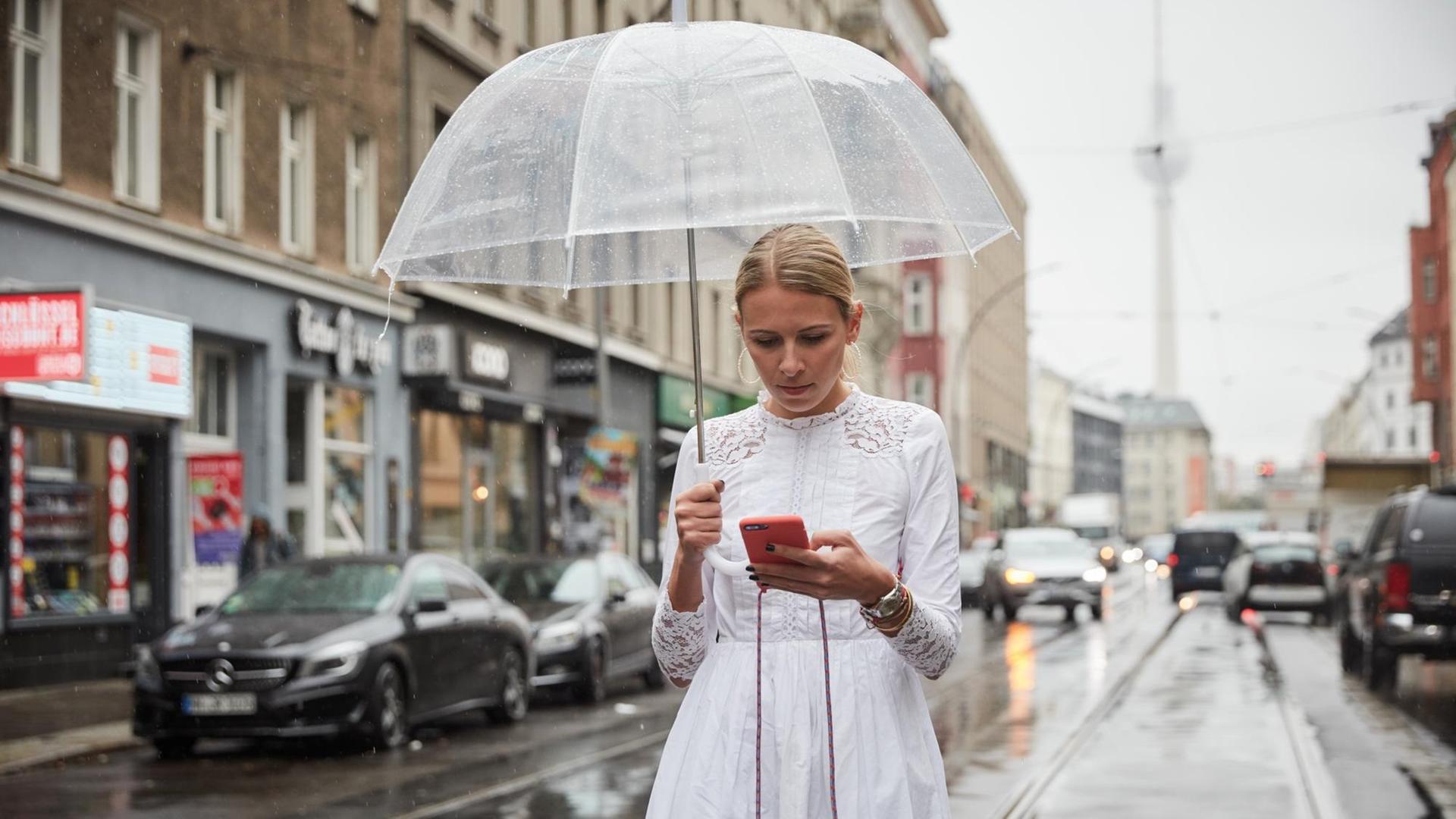 Nena Schink mit einem durchsichtigen Regenschirm und in weißem Kleid in Berlin. Sie tippt auf dem Smartphone. Im Hintergrund ist der Fernsehturm zu erkennen. Nena Schink mit einem durchsichtigen Regenschirm und in weißem Kleid in Berlin. Sie tippt auf dem Smartphone. Im Hintergrund ist der Fernsehturm zu erkennen.