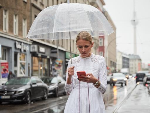 Nena Schink mit einem durchsichtigen Regenschirm und in weißem Kleid in Berlin. Sie tippt auf dem Smartphone. Im Hintergrund ist der Fernsehturm zu erkennen.