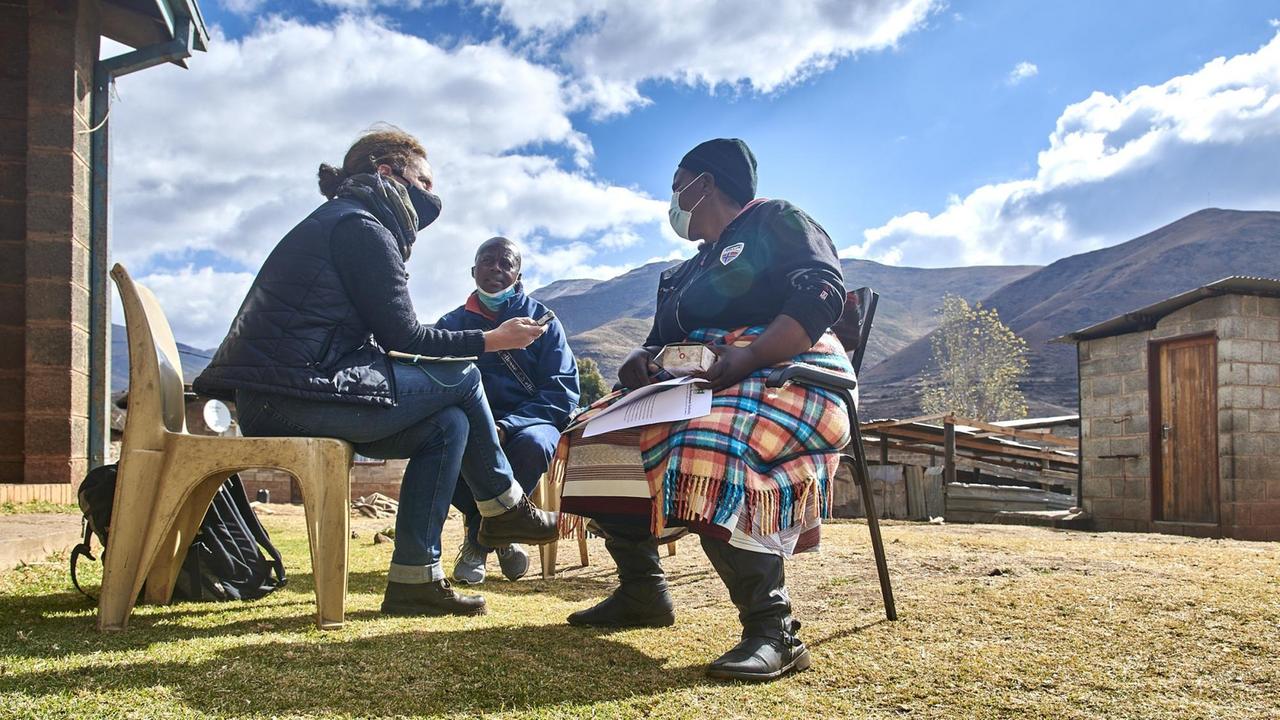 Sie sitzen im Dorf vor einer Hütte auf einfachen Stühlen. Im Hintergrund die Berglandschaft von Lesotho.