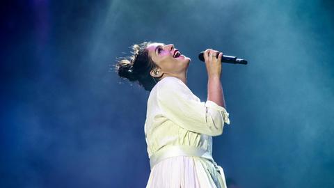 Jessie Ware bei einem Auftritt auf der Bühne: Sie trägt ein weißes Kleid, legt den Kopf in den Nacken und singt in ein Mikrofon, der Hintergrund ist bläulich mit Scheinwerfern.