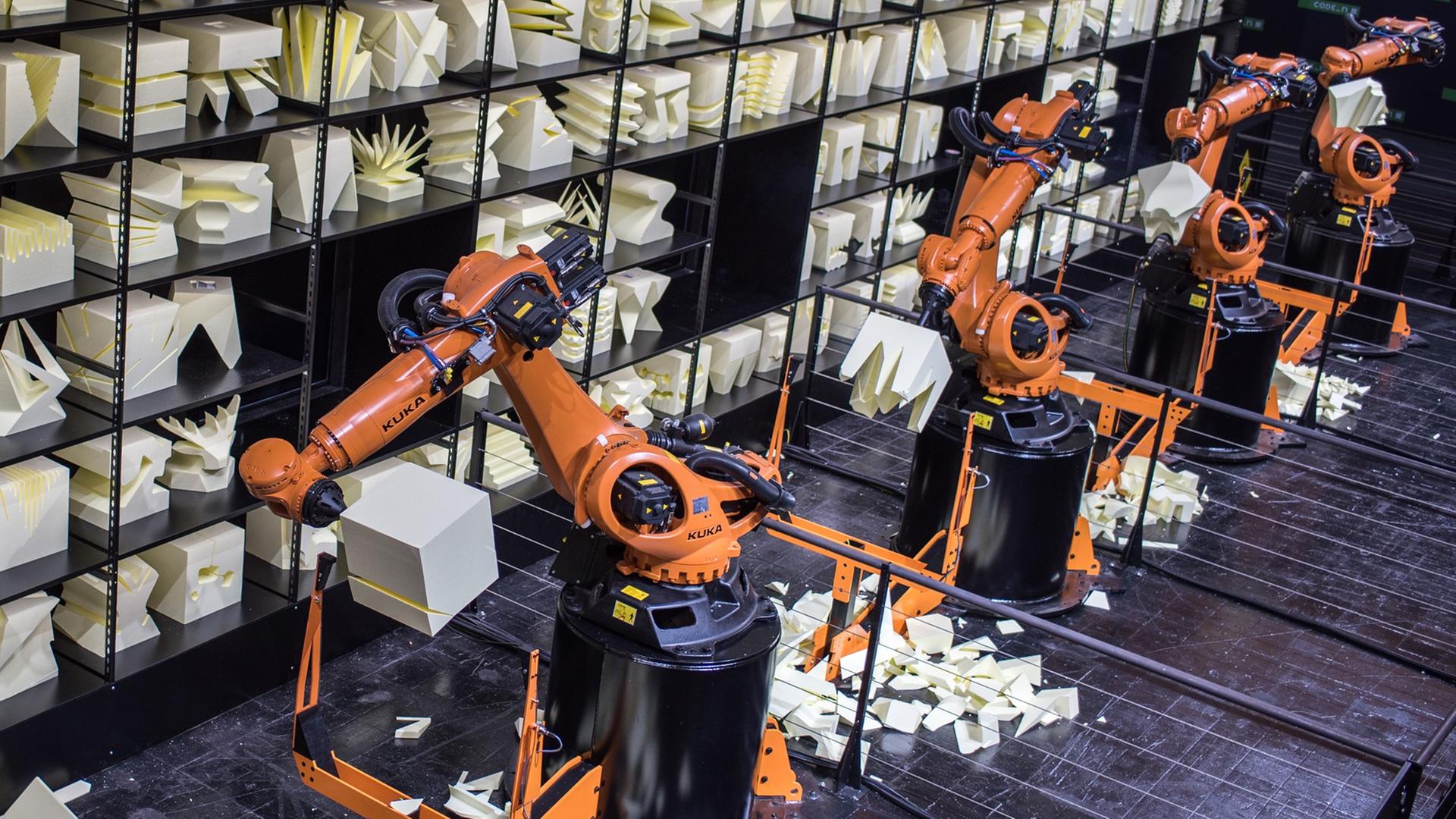 Orangefarbener Produktionsroboterarm, im Hintergrund ein Regal mit Kunstobjekten aus Holz. Orangefarbener Produktionsroboterarm, im Hintergrund ein Regal mit Kunstobjekten aus Holz.