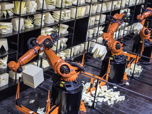 Orangefarbener Produktionsroboterarm, im Hintergrund ein Regal mit Kunstobjekten aus Holz.