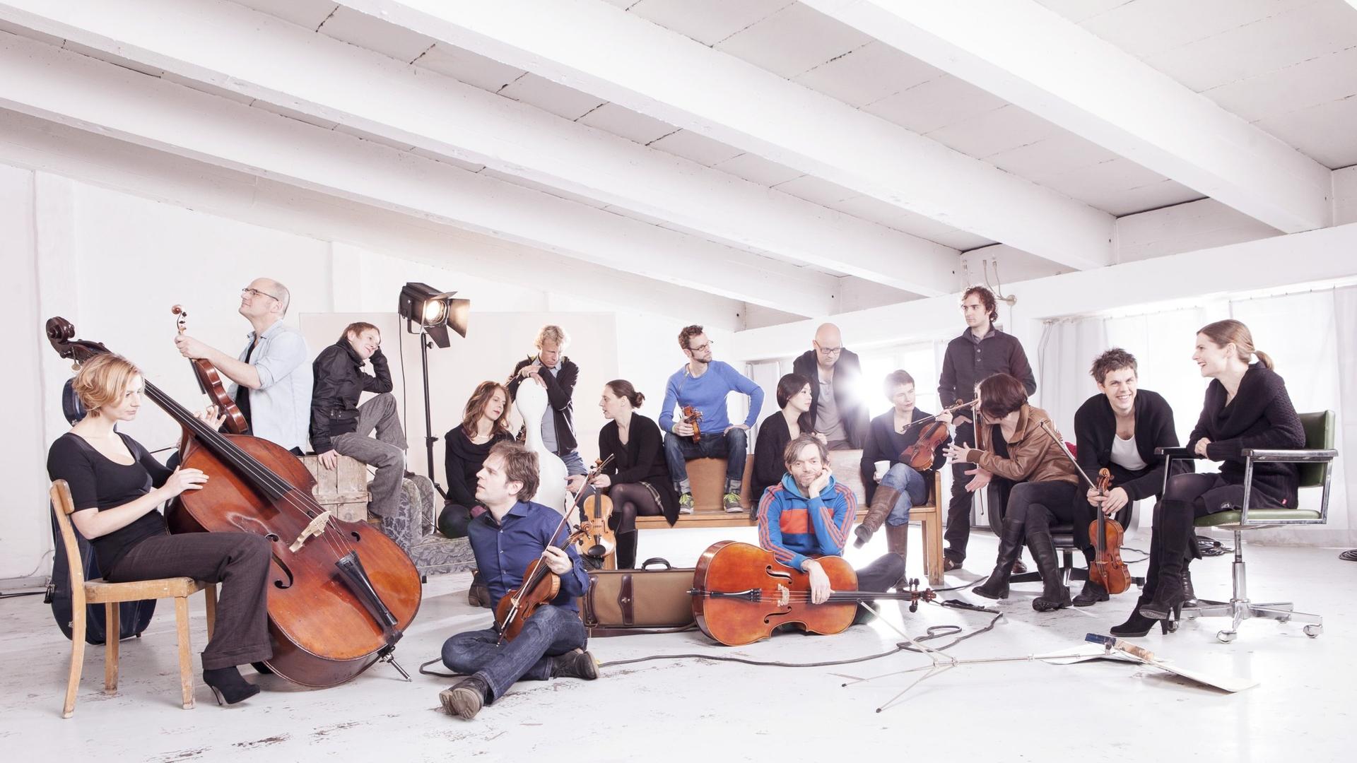 Das Ensemble sitzt und steht mit seinen Instrumenten in einem weißen Raum mit markanter Deckenkonstrukion. Das Ensemble sitzt und steht mit seinen Instrumenten in einem weißen Raum mit markanter Deckenkonstrukion.