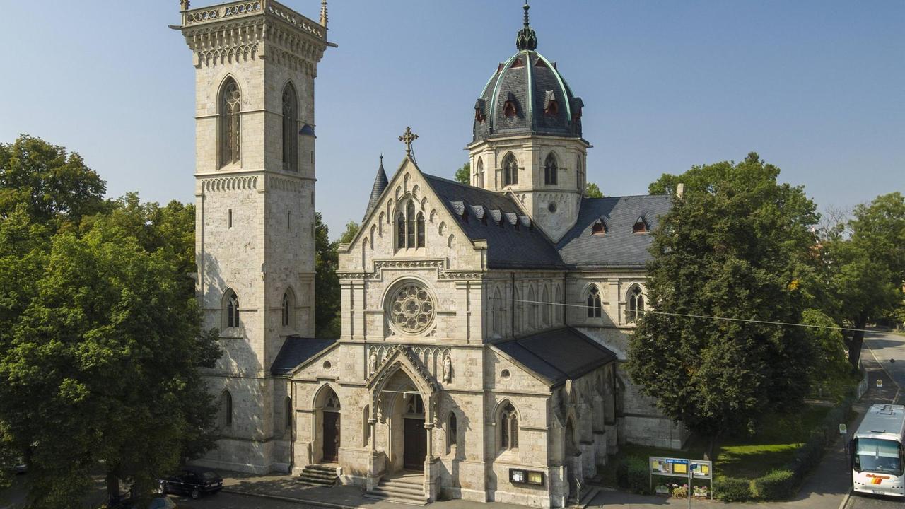 Gesamtansicht der im neugotischen Stil errichteten Herz-Jesu-Kirche in Weimar, die von Bäumen und Autos umgeben ist.