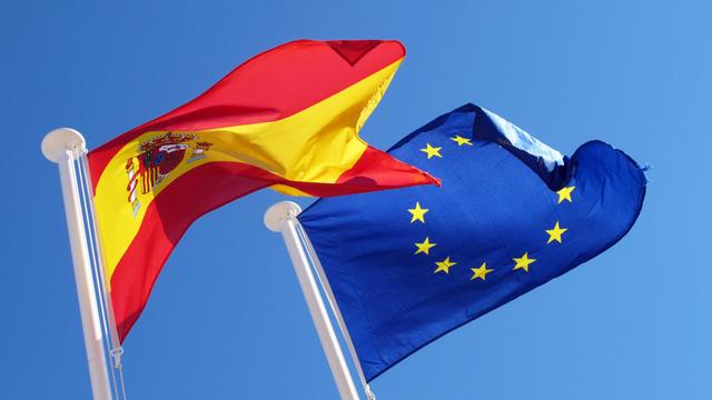Spaniens Flagge und Europafahne wehen vor blauem Himmel im Wind.