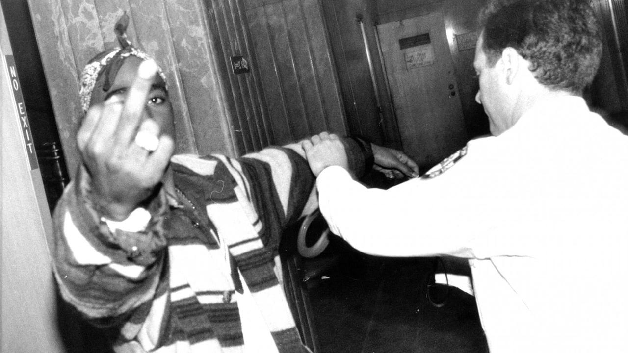 Der Rapper Tupac Shakur 1993 bei einem Termin am Kriminalgericht im New Yorker Stadtteil Manhatten. Tupac guckt in die Kamere und streckt ihr seinen Mittelfinger entgegen. Ein Polizist versucht ihm Handschellen anzulegen.