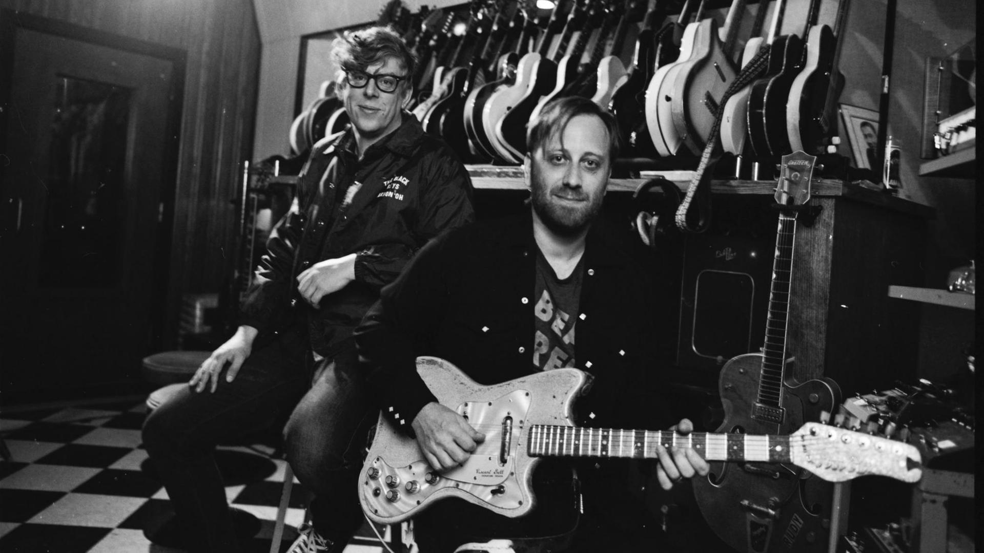 Die beiden Musiker stehen vor einer Wand an der einige Gitarren hängen und schauen in die Kamera. Die beiden Musiker stehen vor einer Wand an der einige Gitarren hängen und schauen in die Kamera.