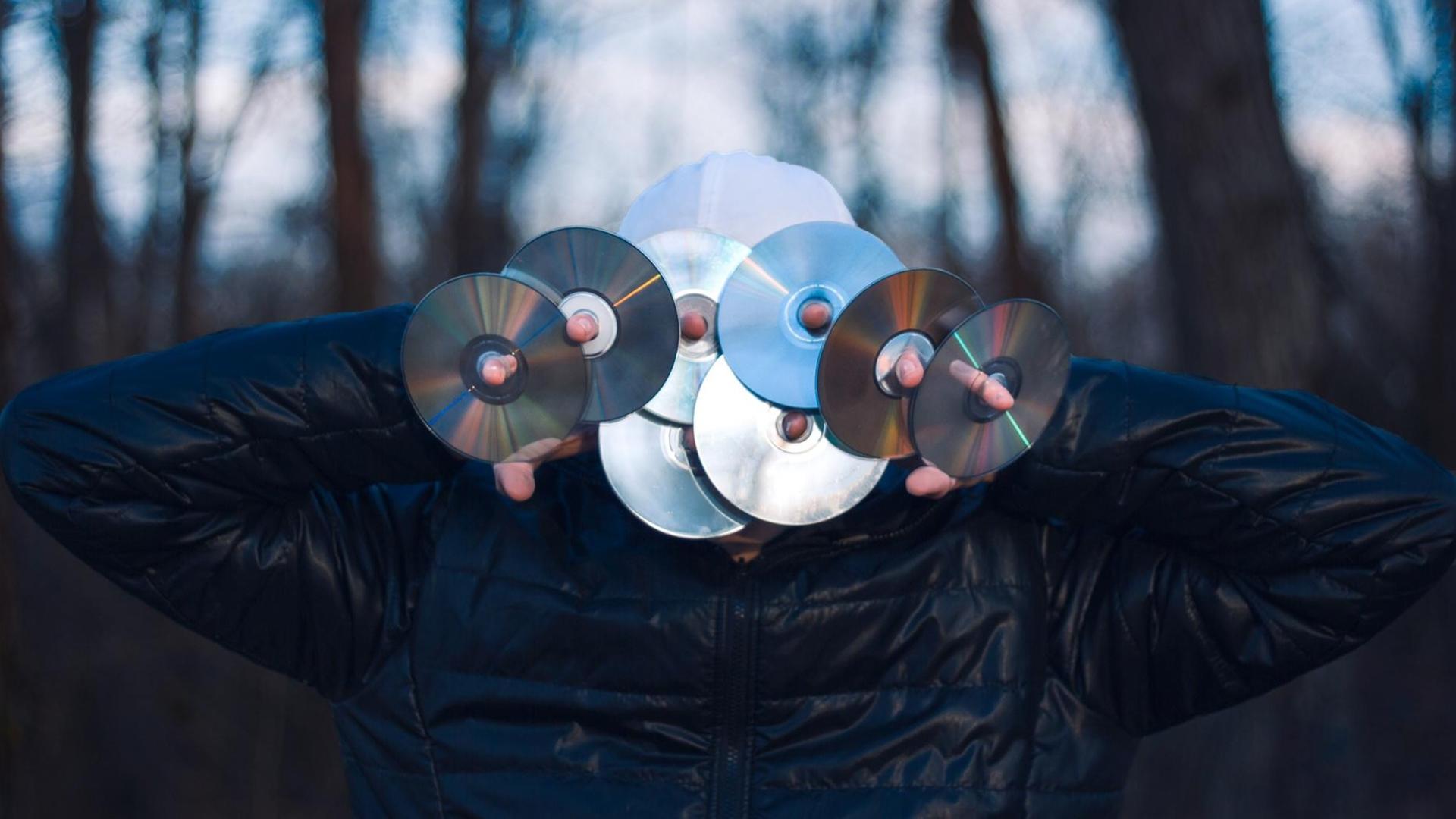 Eine Person hält sich acht CDs vor das Gesicht. Sie steht mitten im Wald. Eine Person hält sich acht CDs vor das Gesicht. Sie steht mitten im Wald.