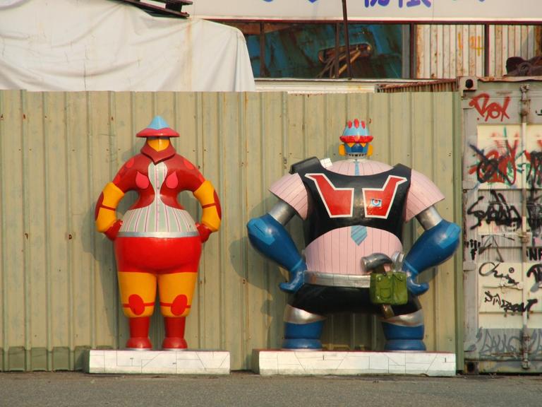 Zwei Roboter-Skulpturen stehen vor einer Baustelle.