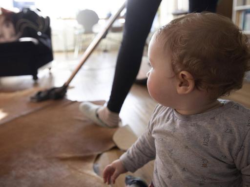 Eine Frau saugt Staub mit einem Staubsauger in ihrem Wohnzimmer. Im Vordergrund sitzt ein Baby auf dem Boden.