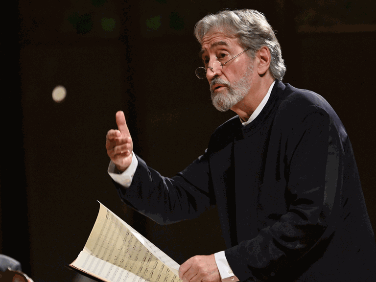 Der weißhaarige Dirigent Jordi Savall trägt schwarzes Sakko und weißes Hemd mit Stehkragen. Er dirigiert mit der rechten Hand und blättert mit der linken die Noten auf dem Pult vor ihm um.
