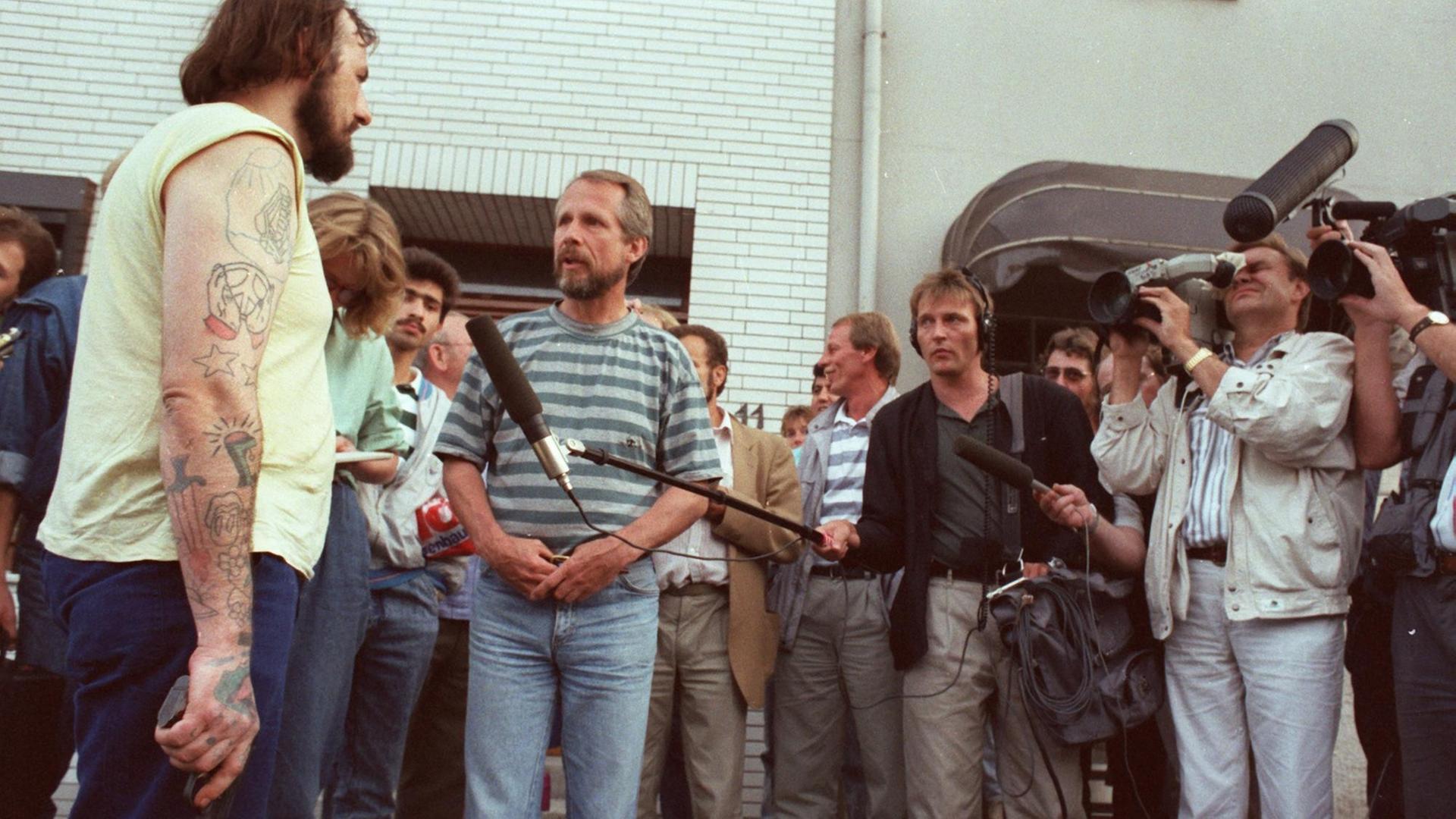 Der Entführer Hans-Jürgen Rösner steht mit einer Pistole in der Hand vor einer Traube von Journalisten, die ihm Mikrofone entgegenstrecken und mit Kameras filmen. Der Entführer Hans-Jürgen Rösner steht mit einer Pistole in der Hand vor einer Traube von Journalisten, die ihm Mikrofone entgegenstrecken und mit Kameras filmen.