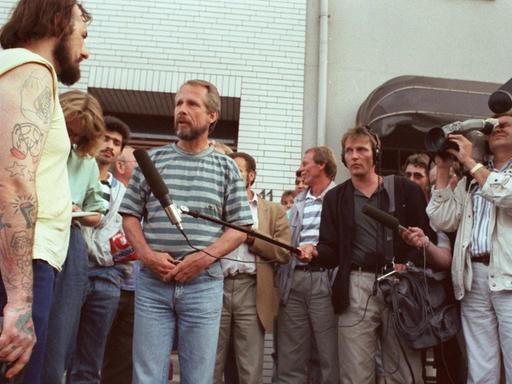Der Entführer Hans-Jürgen Rösner steht mit einer Pistole in der Hand vor einer Traube von Journalisten, die ihm Mikrofone entgegenstrecken und mit Kameras filmen.