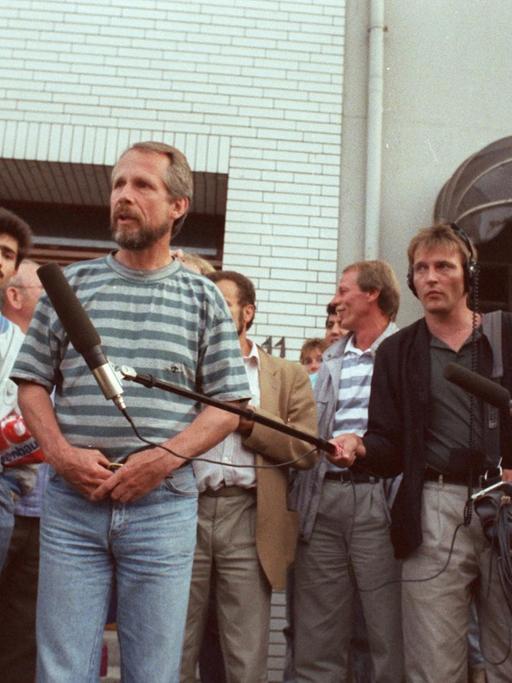 Der Entführer Hans-Jürgen Rösner steht mit einer Pistole in der Hand vor einer Traube von Journalisten, die ihm Mikrofone entgegenstrecken und mit Kameras filmen.