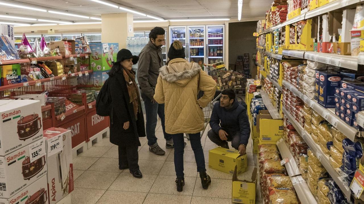 Vier Personen, darunter eine Frau in Schwarz mit Hut, stehen in einem Supermarkt und wählen Produkte aus.