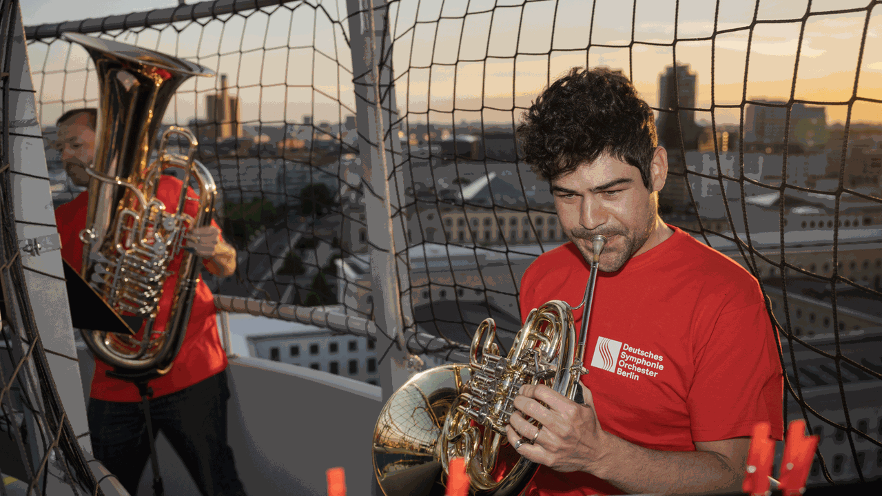Vor einem Ballonkorb-Gitter stehen die Blechbläser Johannes Lipp (Tuba) und Antonio Adriani (Horn) und spielen über den Dächern Berlins. Vor einem Ballonkorb-Gitter stehen die Blechbläser Johannes Lipp (Tuba) und Antonio Adriani (Horn) und spielen über den Dächern Berlins.