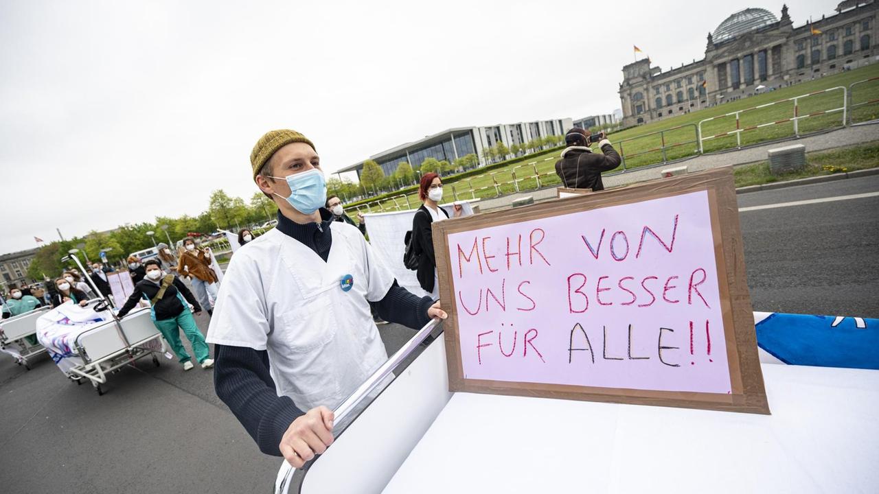 Ein Teilnehmer der Aktion «Der Pflege geht die Luft aus» der Bundespflegekammer und des Berliner Walk of Care geht mit einem Krankenhausbett und einem Schild mit der Aufschrift "Mehr von uns besser für alle" am Reichstagsgebäude vorbei Ein Teilnehmer der Aktion «Der Pflege geht die Luft aus» der Bundespflegekammer und des Berliner Walk of Care geht mit einem Krankenhausbett und einem Schild mit der Aufschrift "Mehr von uns besser für alle" am Reichstagsgebäude vorbei