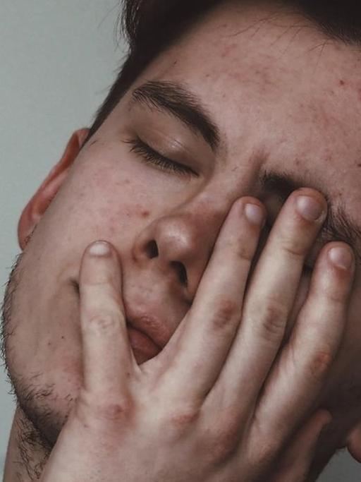 Ein erschöpfter junger Mann legt sein Gesicht mit geschlossenen Augen in seine Hand