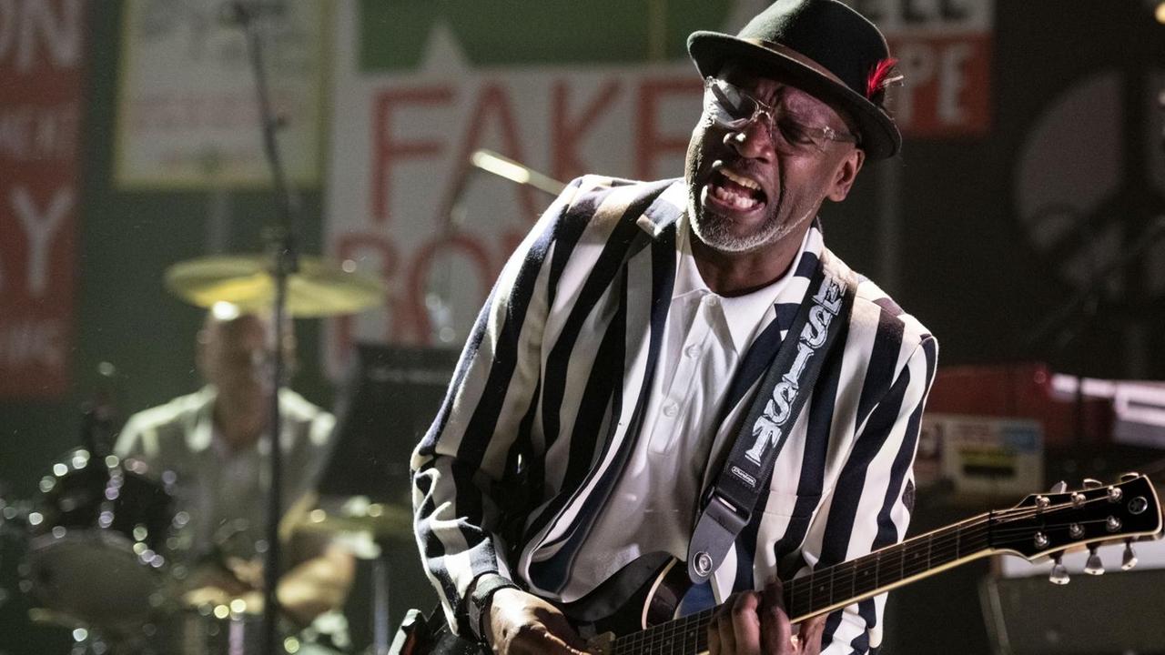 Lynval Golding in einer schwarz-weiß gestreiften Jacke und einer Gitarre. Im Hintergrund sind verschiedene Protestplakate zu erkennen. Lynval Golding in einer schwarz-weiß gestreiften Jacke und einer Gitarre. Im Hintergrund sind verschiedene Protestplakate zu erkennen.