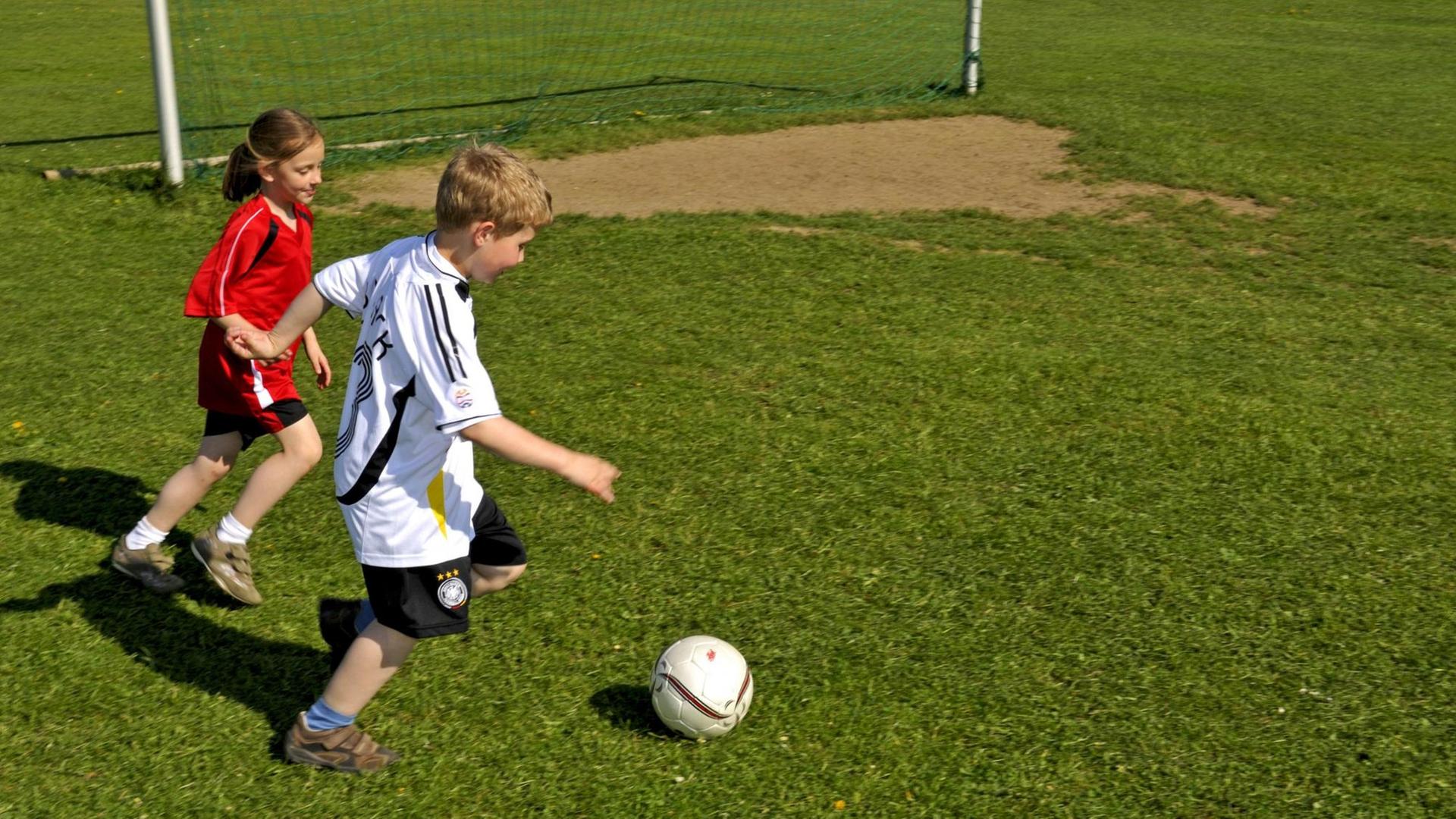 Ein Mädchen, in einem roten Trikot, spielt mit einem Jungen, im weißen Trikot, Fußball.