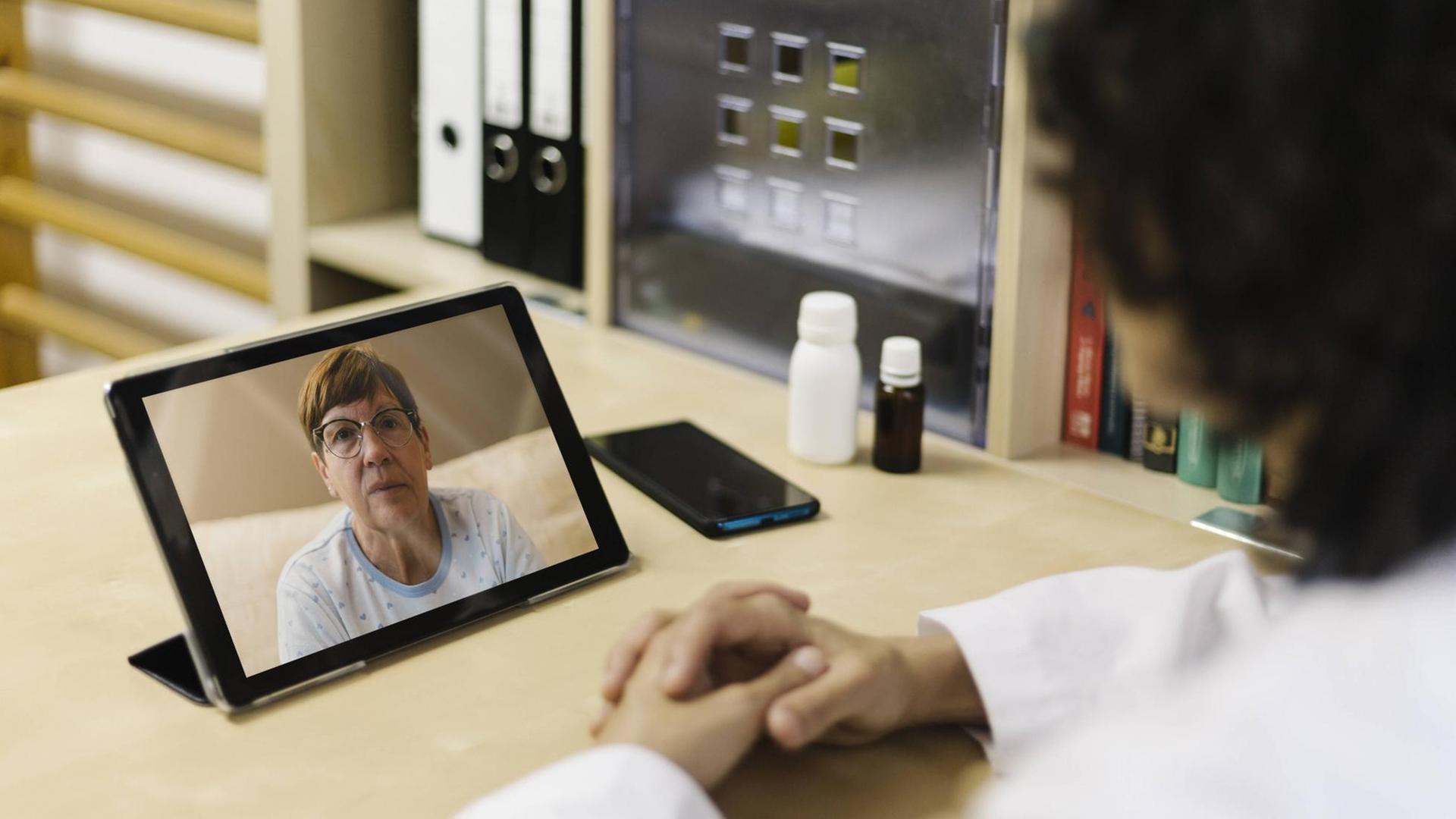 Eine Ärztin sitzt in ihrer Praxis und spricht per Videokonferenz mit einer Patientin, die in ihrem Zuhause ist.
