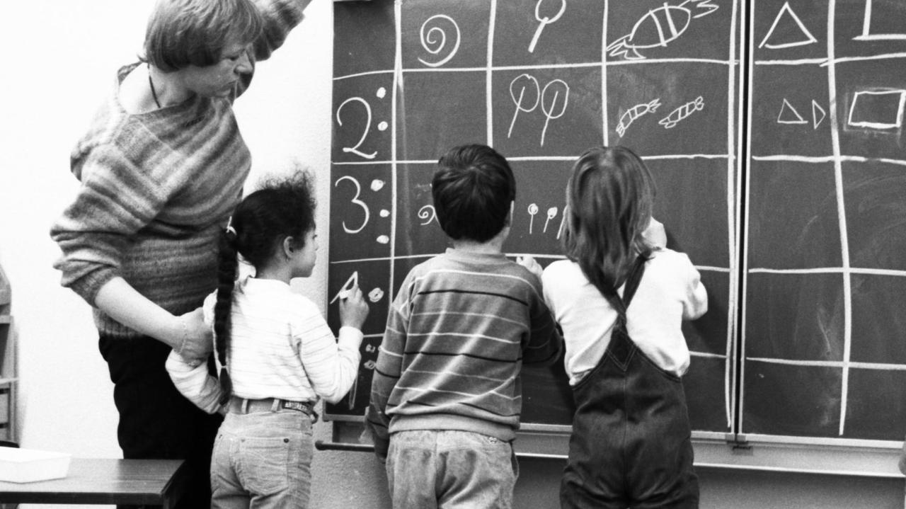 Eine Lehrerin steht mit drei Schülern zusammen an der Tafel und trägt mit Kreide die Lösungen ein.