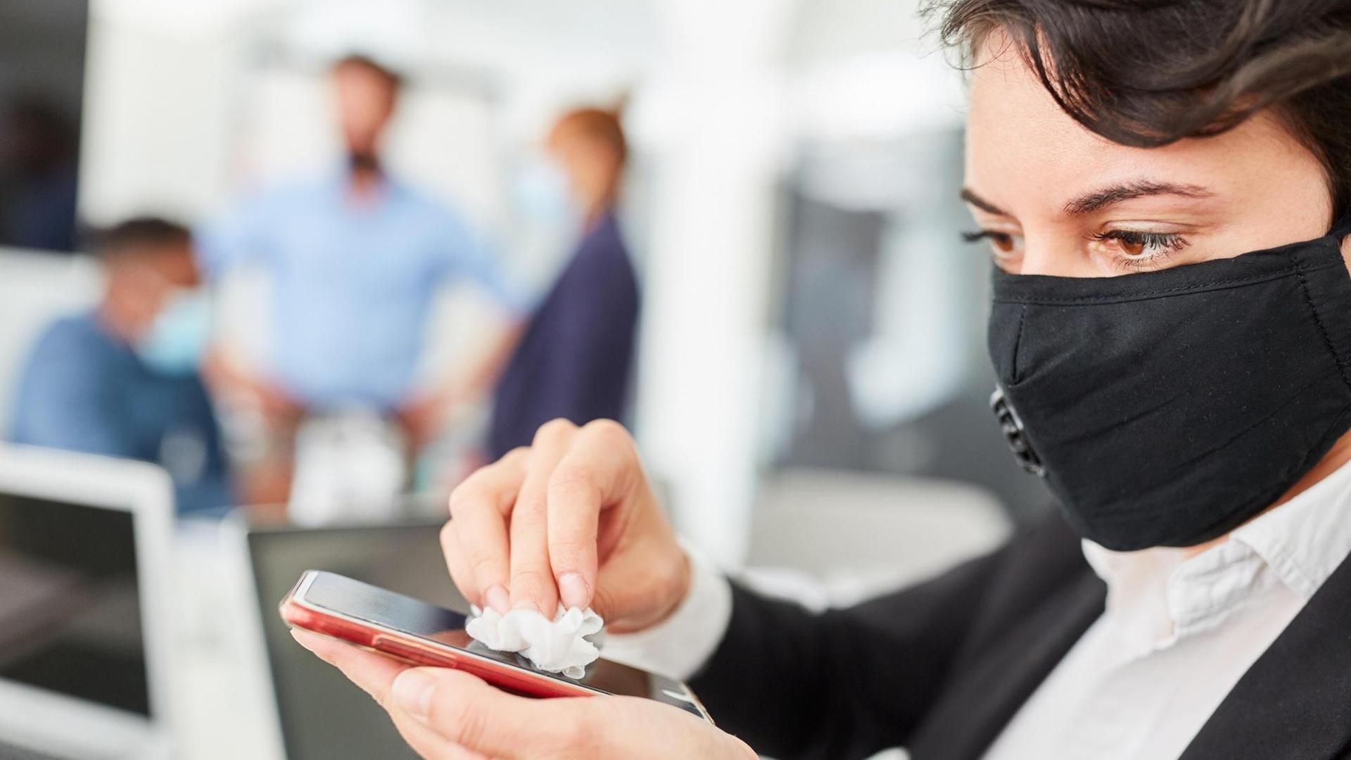 Geschäftsmann mit Maske desinfiziert Smartphone wegen Covid-19 im Büro