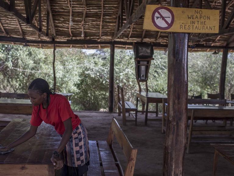 Eine Frau wischt in Kenia über den Tisch eines Restaurants. An einer Pfeiler ist ein Schild mit der Aufschrift "No Weapons in the Restaurant" angebracht.