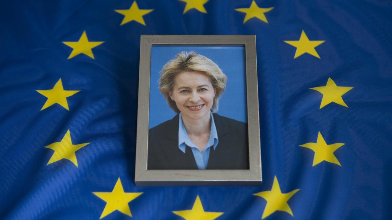 Das Bild zeigt ein gerahmtes Foto von der Politikerin Ursula von der Leyen. Es liegt auf einer blauen Europa-Flagge. Das Bild zeigt ein gerahmtes Foto von der Politikerin Ursula von der Leyen. Es liegt auf einer blauen Europa-Flagge.