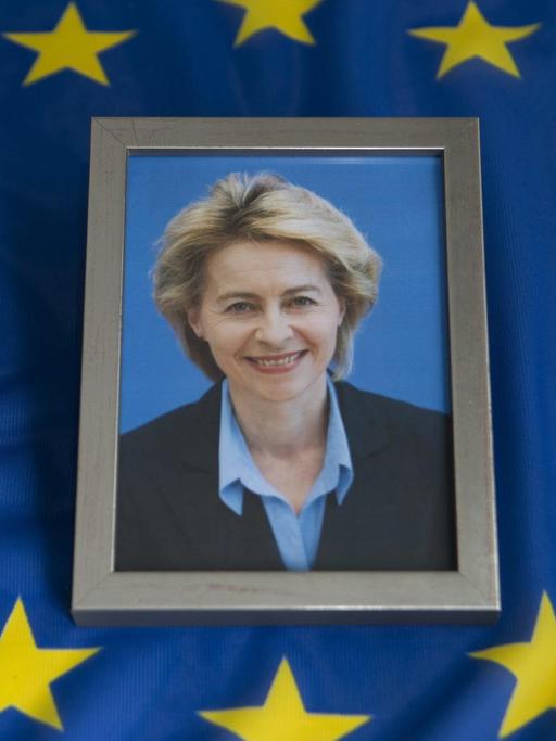 Das Bild zeigt ein gerahmtes Foto von der Politikerin Ursula von der Leyen. Es liegt auf einer blauen Europa-Flagge.