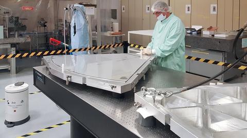 Laborant mit Mundschutz steht an einem Labortisch, auf dem ein etwa zwei Quadratmeter großes Segment des M4-Spiegels des ELT liegt