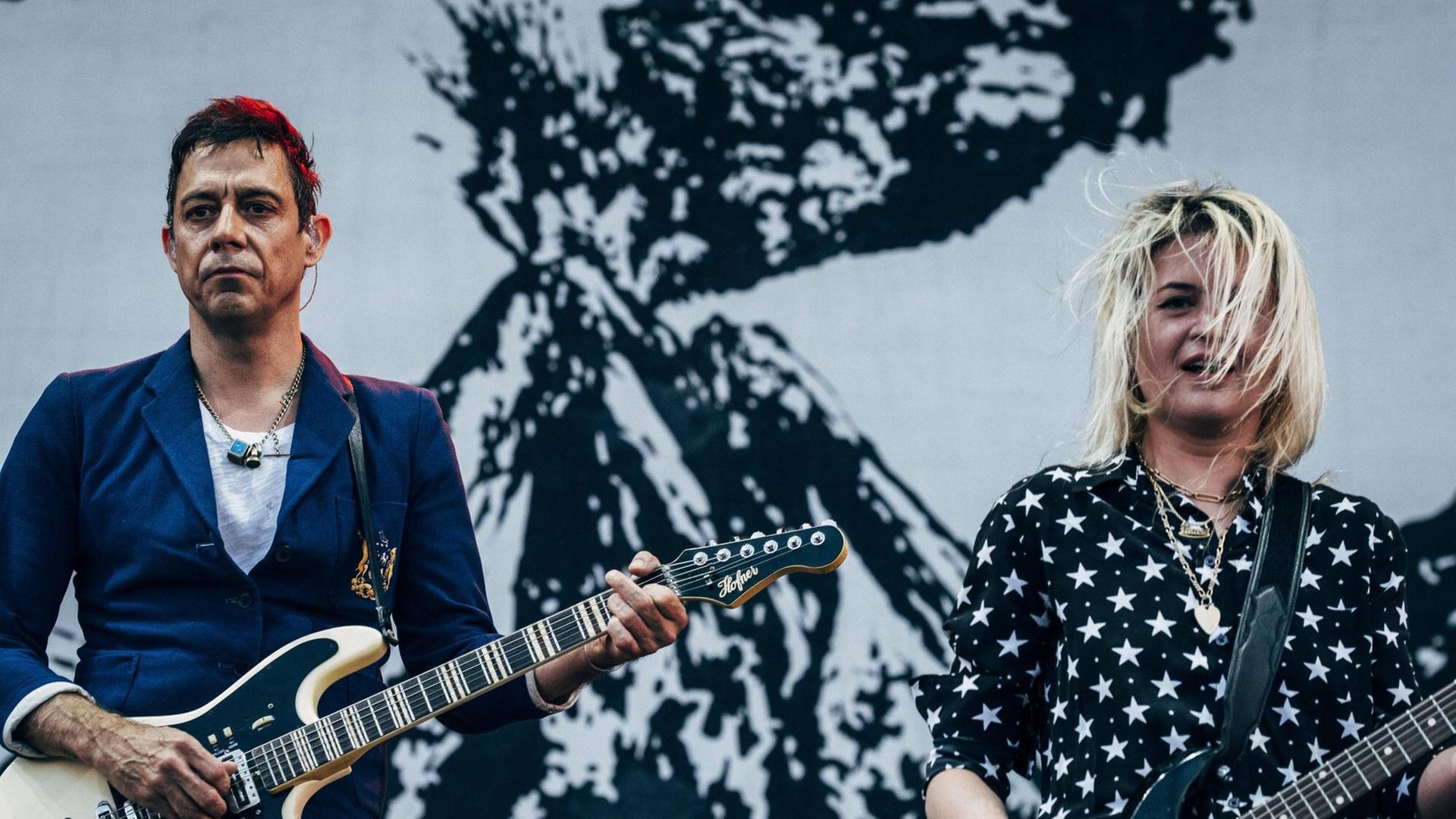 Ein Mann mit kurzen schwarzen Haaren und eine blonde Frau stehen jeweils mit einer Elektro-Gitarre vor dem Bauch auf einer Bühne. Ein Mann mit kurzen schwarzen Haaren und eine blonde Frau stehen jeweils mit einer Elektro-Gitarre vor dem Bauch auf einer Bühne.