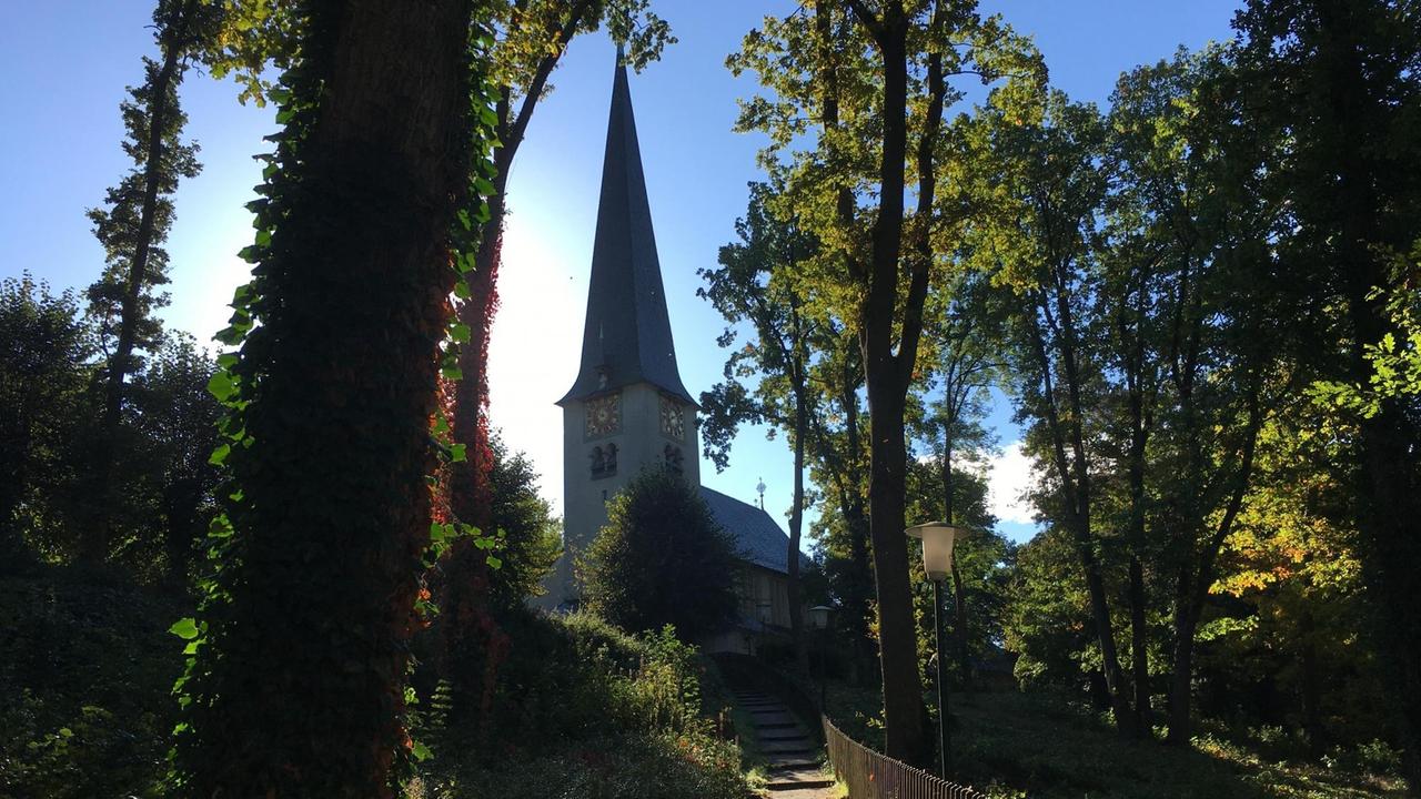 Vor strahlend blauem Himmel erhebt sich eine Kirche mit spitzem Turmdach - im Vordergrund führt ein Weg zwischen Bäumen den Hang hinauf zur Kirche hin. Vor strahlend blauem Himmel erhebt sich eine Kirche mit spitzem Turmdach - im Vordergrund führt ein Weg zwischen Bäumen den Hang hinauf zur Kirche hin.