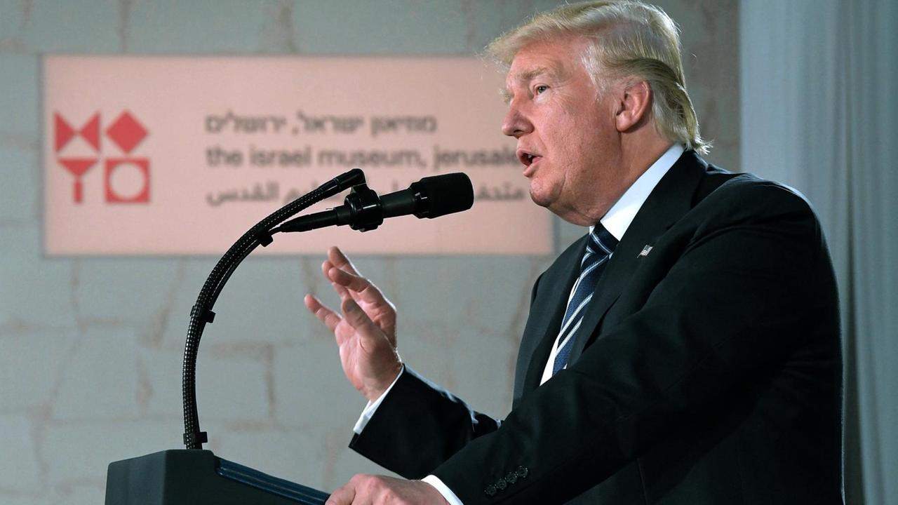 Man sieht Trump redend und gestikulierend von der Seite am Rednerpult. Im Hintergrund an einer Wand ein Schild mit hebräischer Schrift.