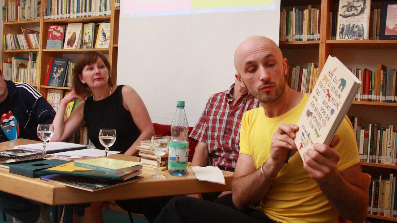 Farbfoto eines jungen Mannes mit Glatze, der ein Buch zeigt bei einer Lesung, es ist der Illustrator Aljoscha Blau Farbfoto eines jungen Mannes mit Glatze, der ein Buch zeigt bei einer Lesung, es ist der Illustrator Aljoscha Blau