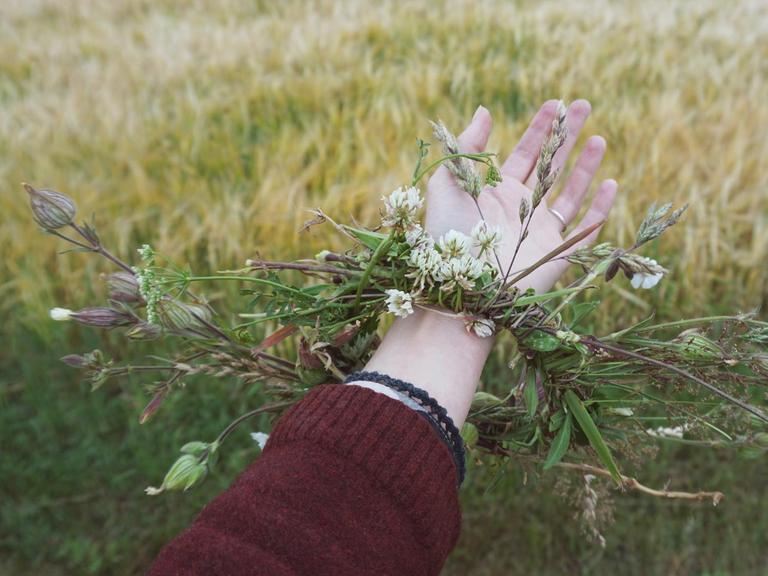 Ein ausgestreckter Arm mit einem Kranz aus wildblühenden Pflanzen um das Handgelenk vor einer Sommerwiese.