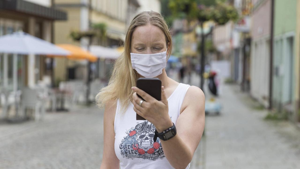 Eine junge Frau mit Mund-Nasen-Behelfs-Schutz schaut in einer Fußgängerzone auf ihr Handy. Eine junge Frau mit Mund-Nasen-Behelfs-Schutz schaut in einer Fußgängerzone auf ihr Handy.