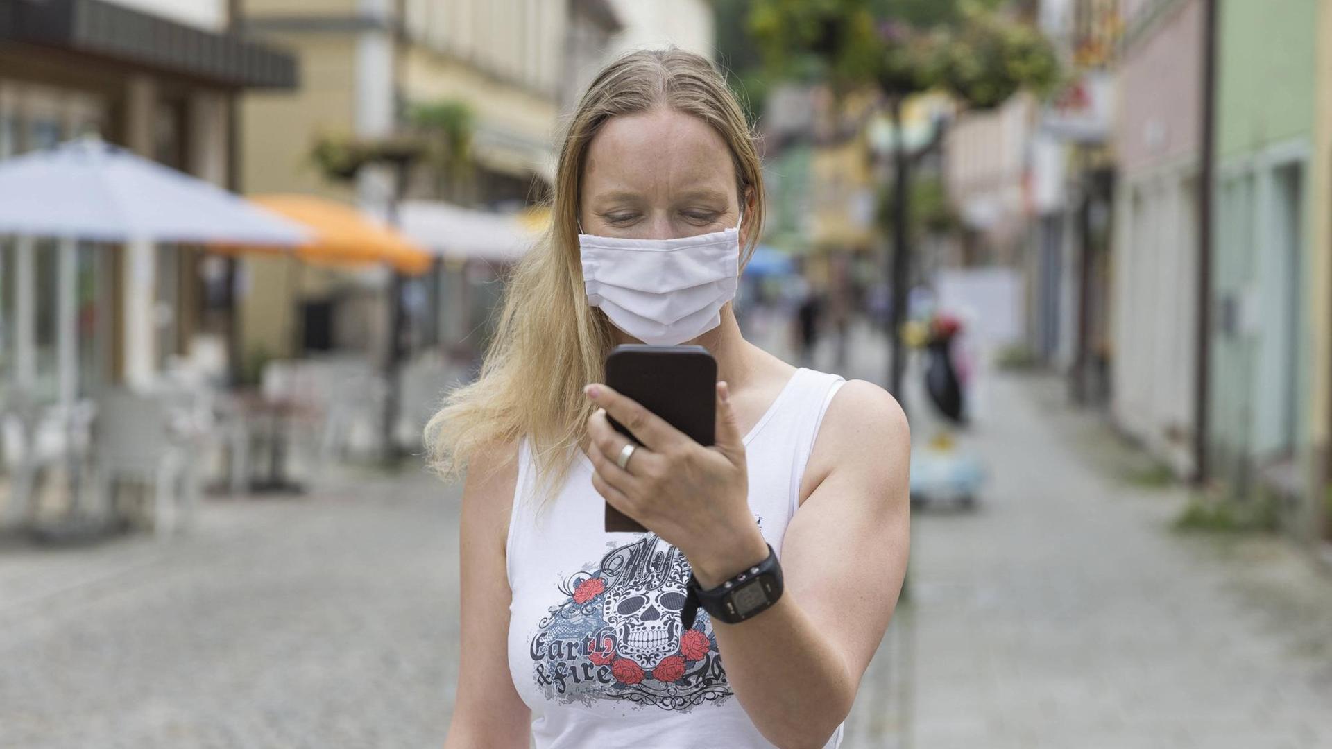 Eine junge Frau mit Mund-Nasen-Behelfs-Schutz schaut in einer Fußgängerzone auf ihr Handy. Eine junge Frau mit Mund-Nasen-Behelfs-Schutz schaut in einer Fußgängerzone auf ihr Handy.
