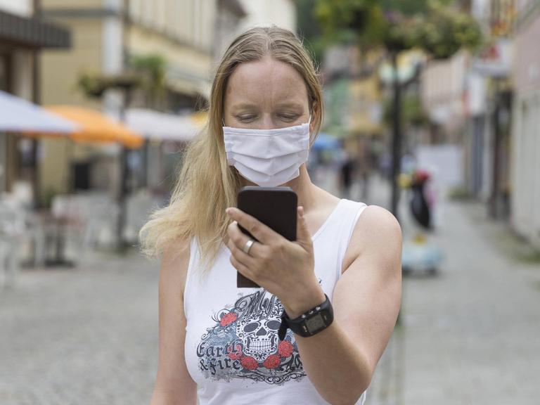 Eine junge Frau mit Mund-Nasen-Behelfs-Schutz schaut in einer Fußgängerzone auf ihr Handy.