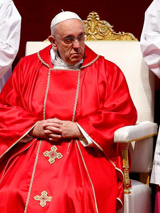 Papst Franziskus sitzt im roten Gewand auf einem weißen Stuhl, rechts und links neben ihm zwei Messdiener in weißen Gewändern mit gefalteten Händen