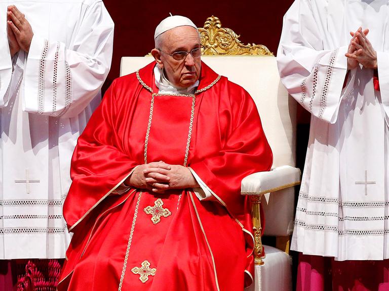 Papst Franziskus sitzt im roten Gewand auf einem weißen Stuhl, rechts und links neben ihm zwei Messdiener in weißen Gewändern mit gefalteten Händen
