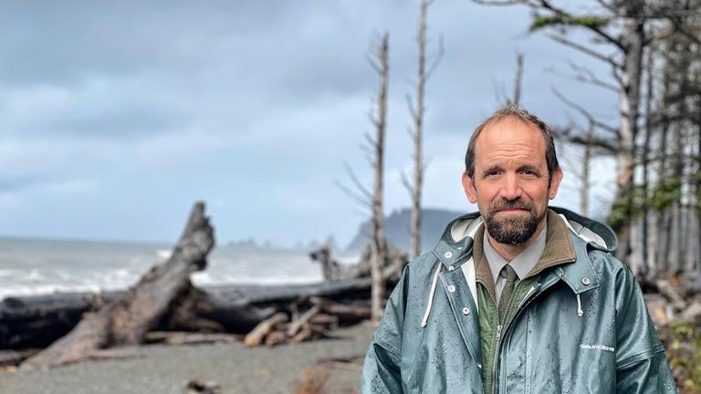 Ein Mann mit Bart und grüner Regenjacke steht an einem Strand mit Treibholz. Ein Mann mit Bart und grüner Regenjacke steht an einem Strand mit Treibholz.