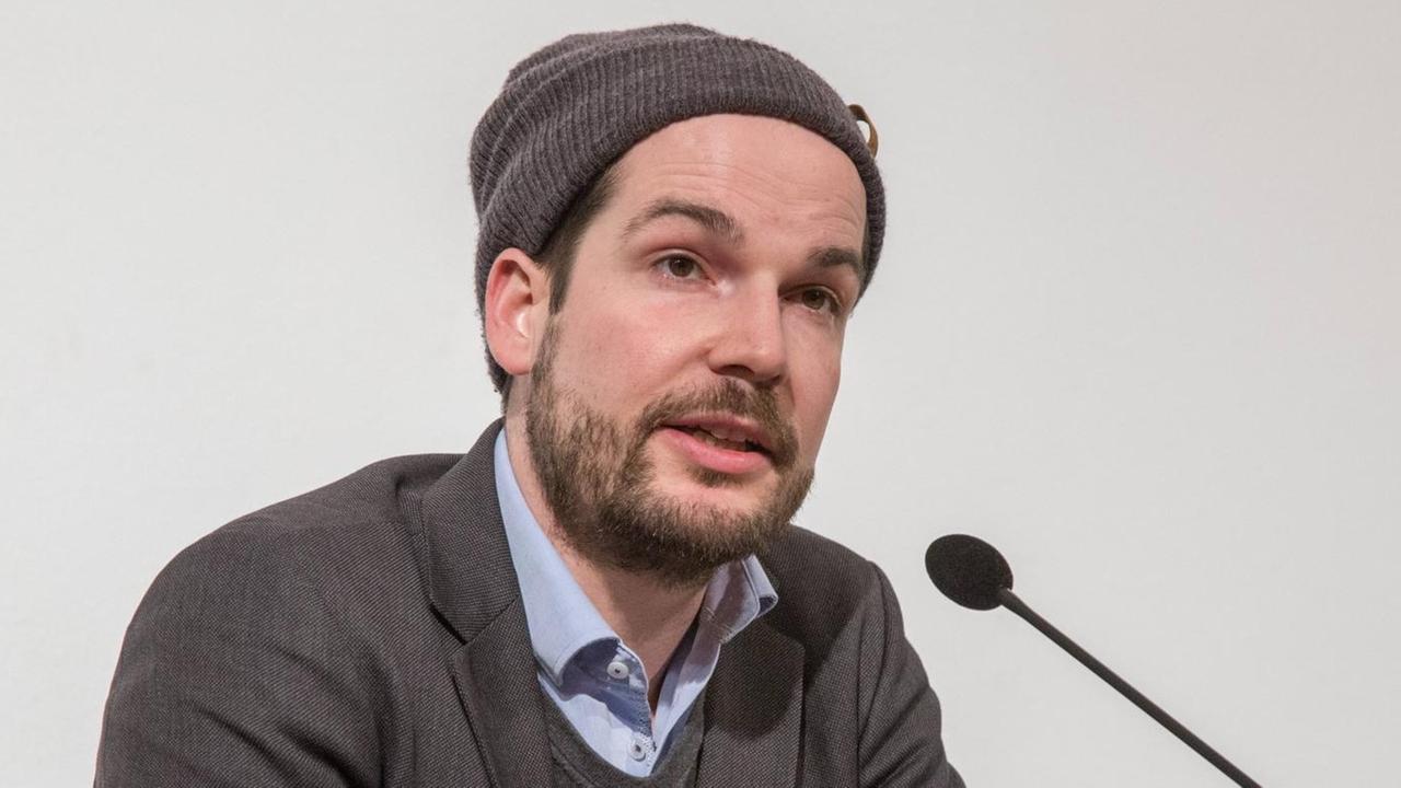 Max Czollek mit Hemd, Pullover und Mütze vor einem Mikrofon auf einer Bühne. Max Czollek mit Hemd, Pullover und Mütze vor einem Mikrofon auf einer Bühne.