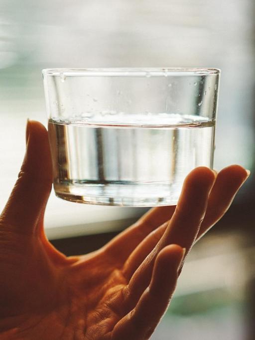 Eine Hand hält ein Glas Wasser. Im Hintergrund ist ein Fenster zu sehen.