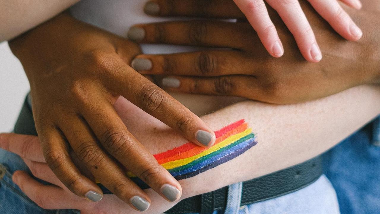 Vier Hände, zwei dunkle und zwei helle, halten sich in einer Umarmung. Auf einem Arm ist ein Regenbogen aufgemalt. Vier Hände, zwei dunkle und zwei helle, halten sich in einer Umarmung. Auf einem Arm ist ein Regenbogen aufgemalt.
