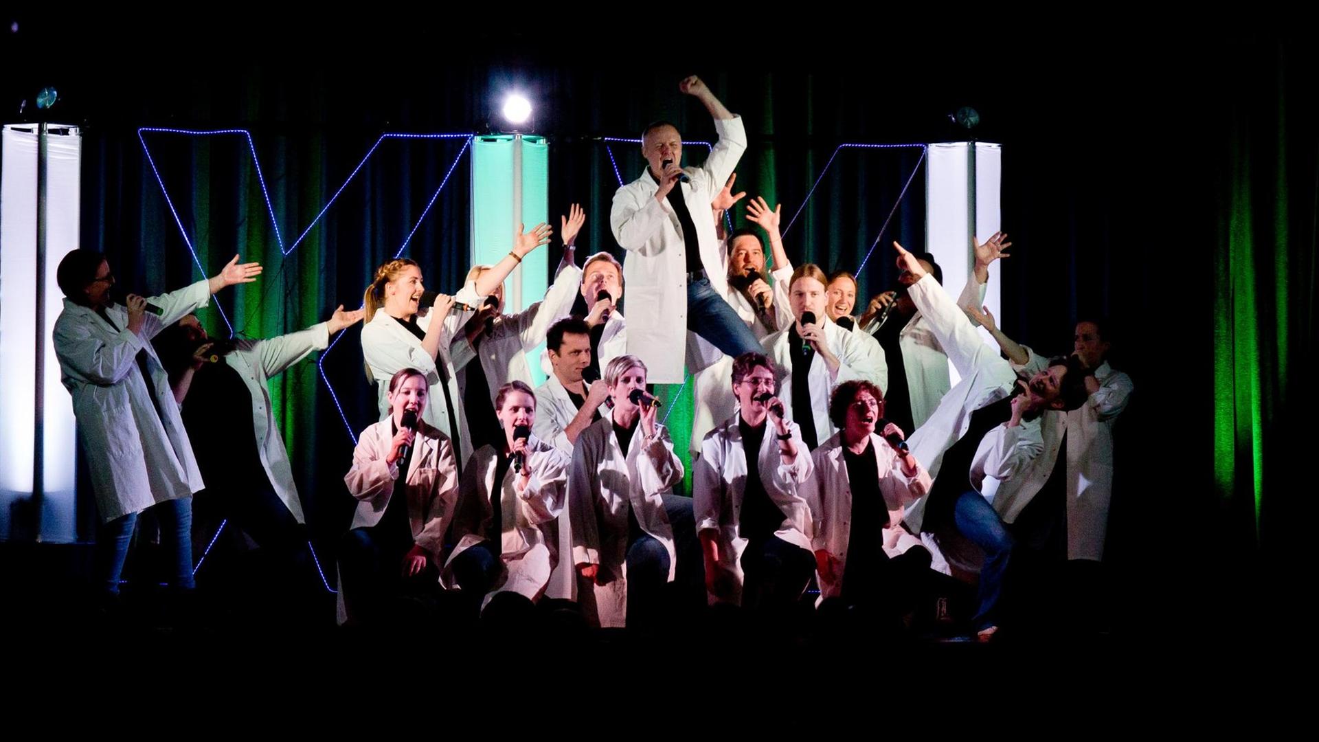 Die Sänger des Chores tragen bei einem Konzert auf der Bühne weiße Kittel. Jeder hält ein Mikrofon in der Hand.